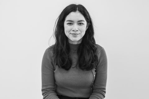 A black and white photo of Ashleigh Vissel with long, dark hair, wearing a ribbed, long-sleeve sweater. They are sitting and facing the camera with a slight smile against a plain background.