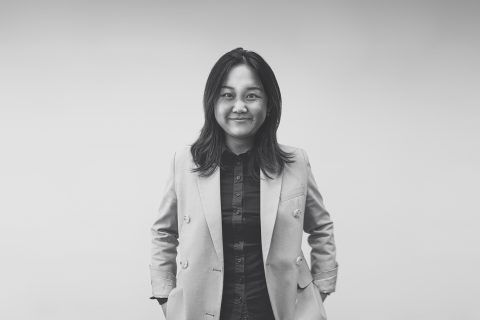 A black-and-white portrait of JJ Zhou with shoulder-length hair, wearing a light-colored blazer and a dark shirt. They are standing against a plain background, smiling slightly and looking directly at the camera with their hands casually in their pockets.