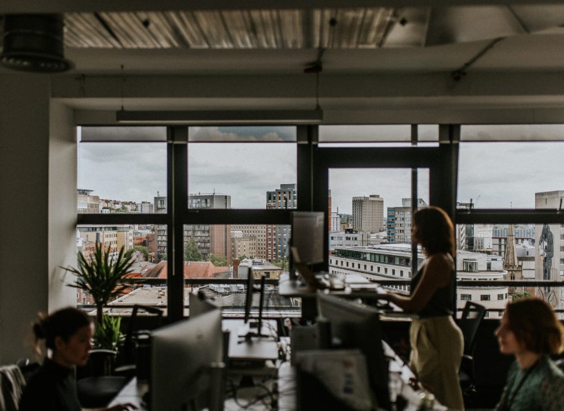 A modern office space with large windows offering a view of city buildings. In the foreground, several desks with computers are seen, and three people are working, with one standing and two sitting. Plants and office equipment are also present.