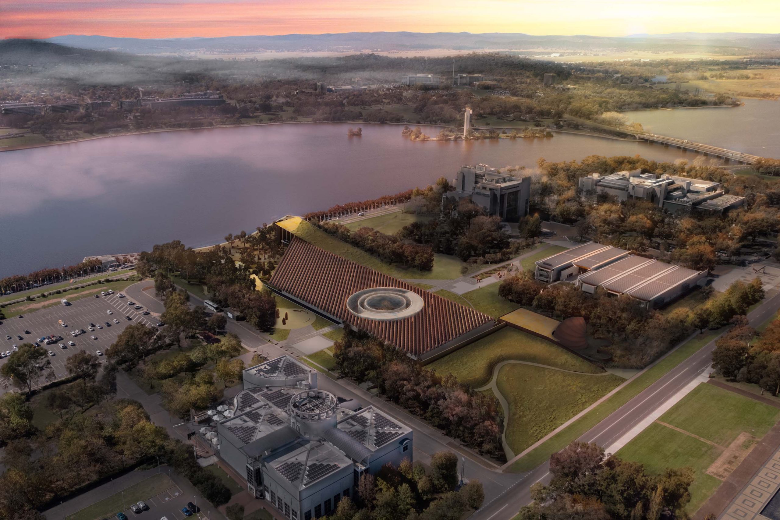 Aerial view of the Ngurra Cultural Precinct's large, pyramid-shaped building with a central circular feature near a waterbody at sunset. Surrounding the pyramid are additional modern structures, parking lots, roads, and trees. The sky is colorful with hues of pink, orange, and yellow.