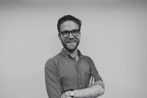 A man with glasses, a beard, and short hair, wearing a long-sleeved button-up shirt and a watch, stands against a plain background. Smiling with his arms crossed, Robert Snelling exudes confidence in this black-and-white image.