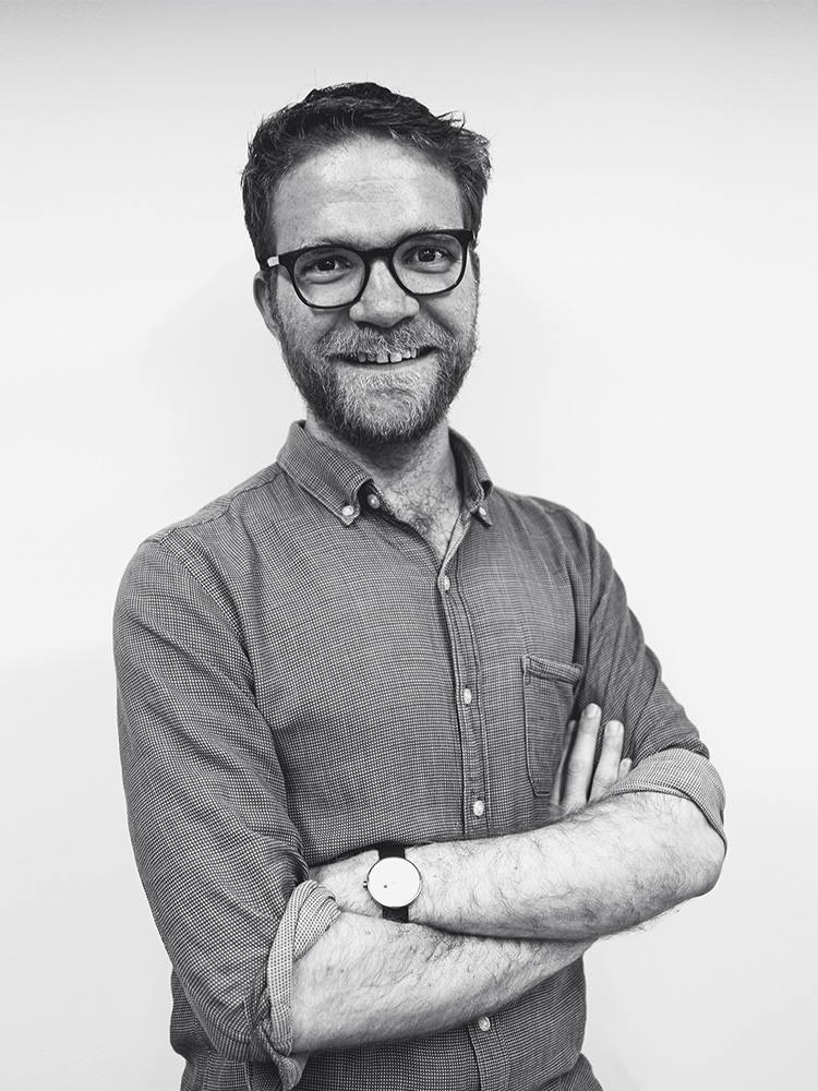 Robert Snelling, a man with glasses, a beard, and a smiling expression, stands against a plain background with his arms crossed. He is wearing a button-up shirt and a wristwatch, and the image is in black and white.
