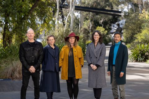 Five people stand side by side outdoors on a path in the National Sculpture Garden, surrounded by greenery. They are wearing various outfits, with one person in a red hat and another in a blue turtleneck. They all face the camera, smiling subtly. Trees and shrubs are visible in the background of this design competition entry.