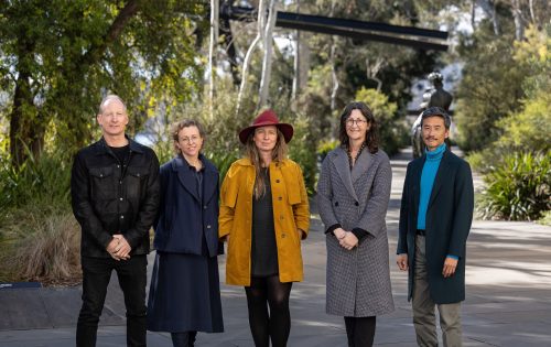 Five people stand side by side outdoors on a path in the National Sculpture Garden, surrounded by greenery. They are wearing various outfits, with one person in a red hat and another in a blue turtleneck. They all face the camera, smiling subtly. Trees and shrubs are visible in the background of this design competition entry.