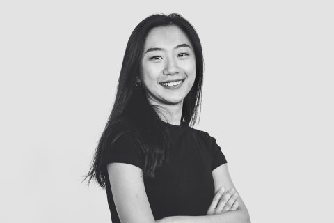 A black and white photo of Tiange Wu, a young woman with long, dark hair, smiling confidently. She is wearing a short-sleeved, dark-colored shirt and stands with her arms crossed against a plain background.