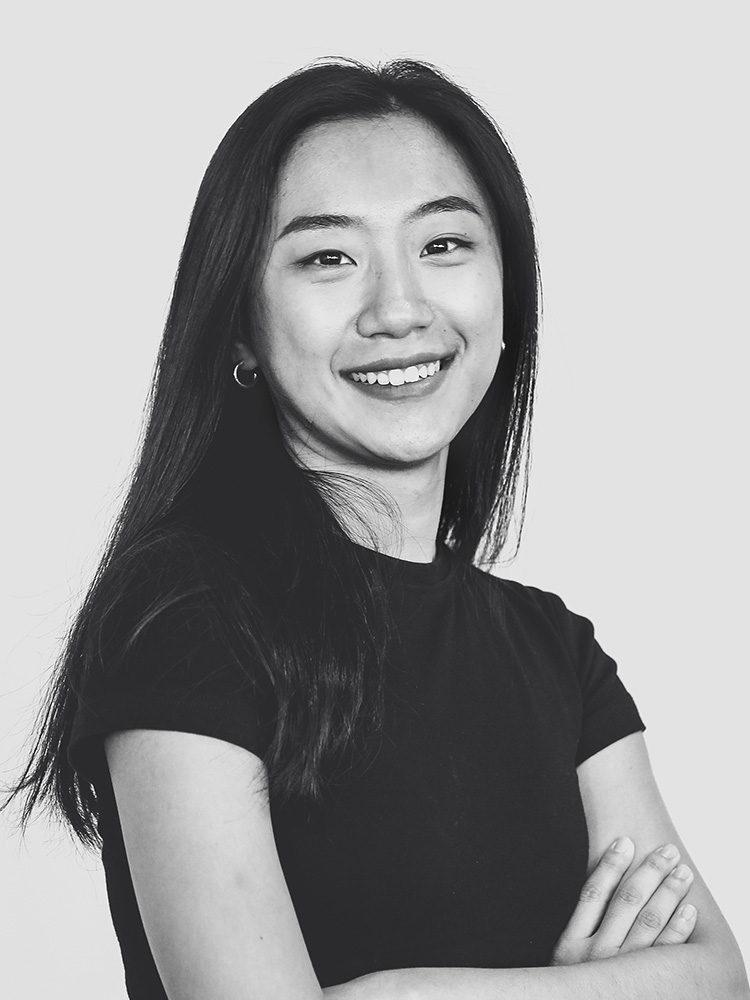 A black and white portrait of Tiange Wu, a person with long, straight hair smiling towards the camera. They are wearing a short-sleeved, dark-colored top and have their arms crossed. The background is plain and light-colored.