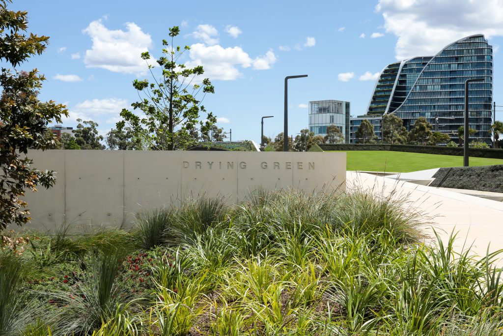 Modern park with lush greenery and a "Drying Green" sign on a concrete wall. Contemporary high-rise buildings with glass facades rise in the background under a bright blue sky with scattered clouds. This award-winning space emphasizes sustainability, featuring well-maintained pathways and streetlights.