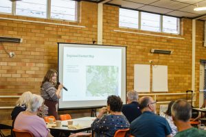 A woman, Remy Ellis, is presenting to a small group of people in a classroom setting. She stands by a projector screen displaying a "Regional Context Map" featuring Bellingen and Dorrigo. The audience members are seated at tables, listening attentively to her insights on Sustainable Master Plans. The room has brick walls and large windows.