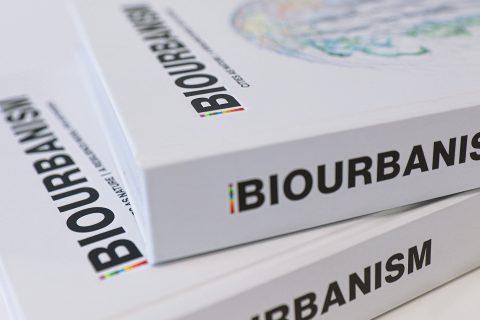 Three white books are stacked, each titled "Biourbanism" with a subtitle, "Cities as Nature." The word "BIOURBANISM" is prominently displayed along the spines of the books. The focus is on the book titles with a partially visible, colorful illustration on the cover, emphasizing design models for climate resilient cities.