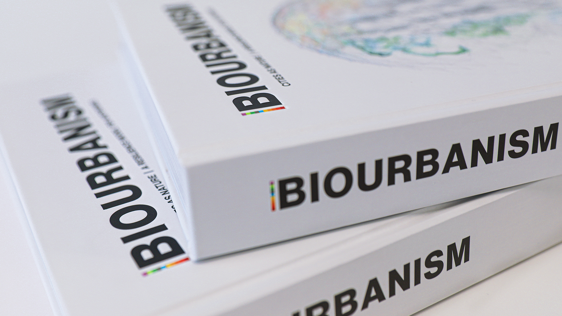 Three white books are stacked, each titled "Biourbanism" with a subtitle, "Cities as Nature." The word "BIOURBANISM" is prominently displayed along the spines of the books. The focus is on the book titles with a partially visible, colorful illustration on the cover, emphasizing design models for climate resilient cities.