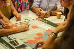 A group of people gather around a table in Bellingen, engaging in a collaborative discussion. They are examining a large map annotated with various colored sticky notes, outlining sustainable master plans. One person points at the map, and another holds a coffee cup.