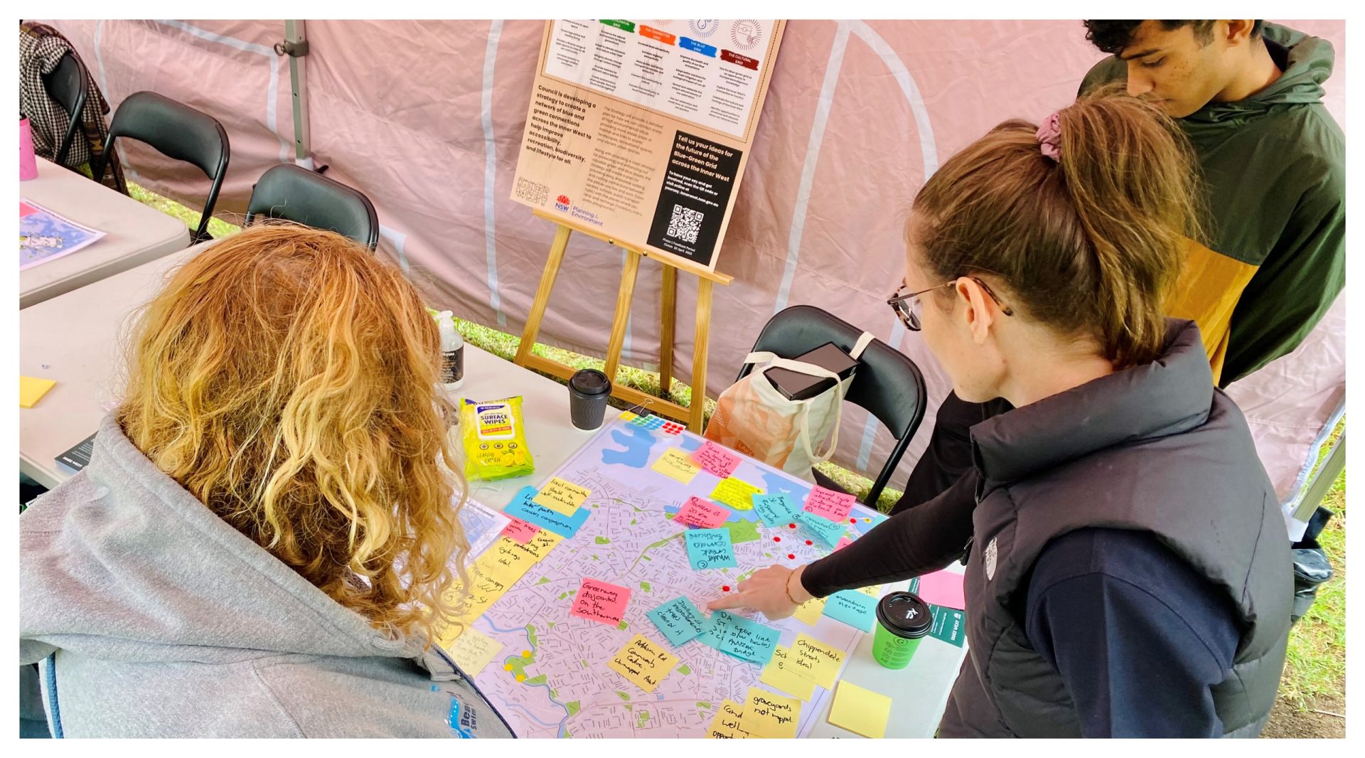 Three people are gathered around a table covered with a large map, colorful sticky notes, and pens. One person, wearing glasses and a vest, points at the map. An informational poster about the Inner West Blue-Green Grid Strategy is displayed on an easel in the background. Cups of coffee are on the table.