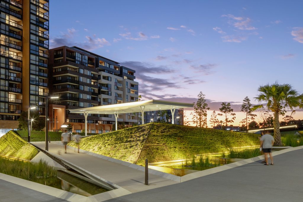 Modern apartment buildings at dusk with geometric landscaping, a canopy structure, and trees in the foreground. People walk along the lit concrete pathways of this sustainability-focused community while the sky displays a colorful sunset with hues of orange, purple, and blue.