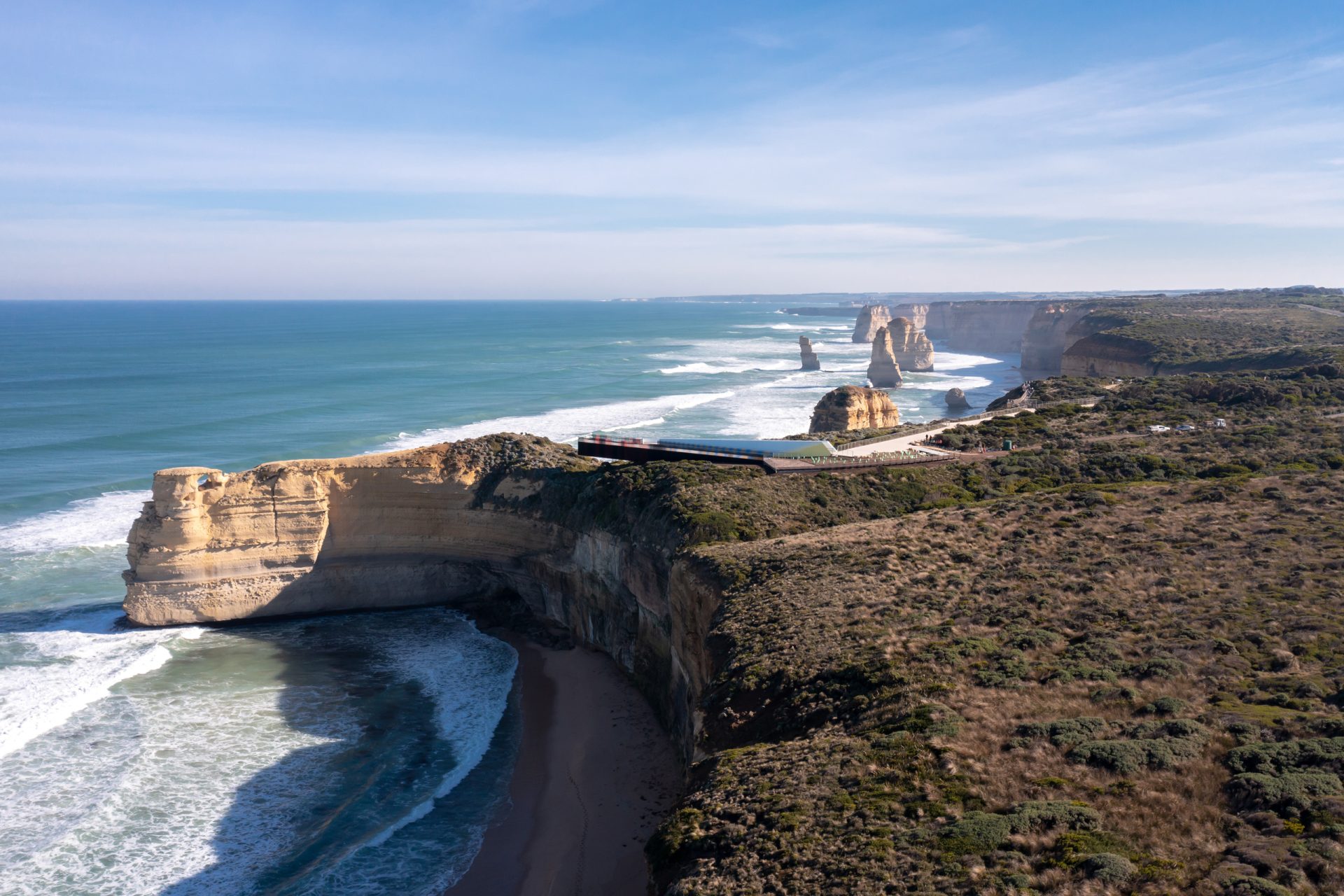 A scenic aerial view of the Twelve Apostles along the Great Ocean Road in Australia. This iconic tourist attraction showcases rugged limestone stacks rising from the ocean, bordered by cliffs and lush greenery. Waves crash against the shore under a clear blue sky, offering a breathtaking vista from every lookout.