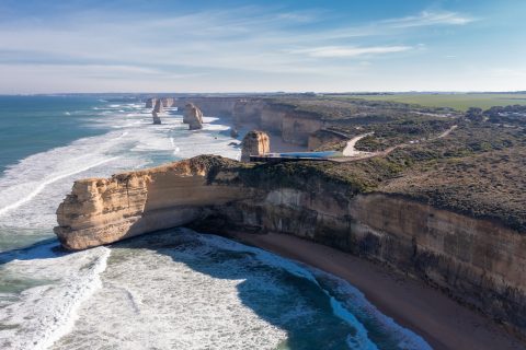 Aerial view of the Twelve Apostles, limestone stacks off the shore of Port Campbell National Park along the Great Ocean Road in Victoria, Australia. From a scenic lookout, witness waves crashing below as these formations majestically rise from the Southern Ocean with a coastal road winding in the background.