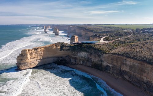 Aerial view of the Twelve Apostles, limestone stacks off the shore of Port Campbell National Park along the Great Ocean Road in Victoria, Australia. From a scenic lookout, witness waves crashing below as these formations majestically rise from the Southern Ocean with a coastal road winding in the background.