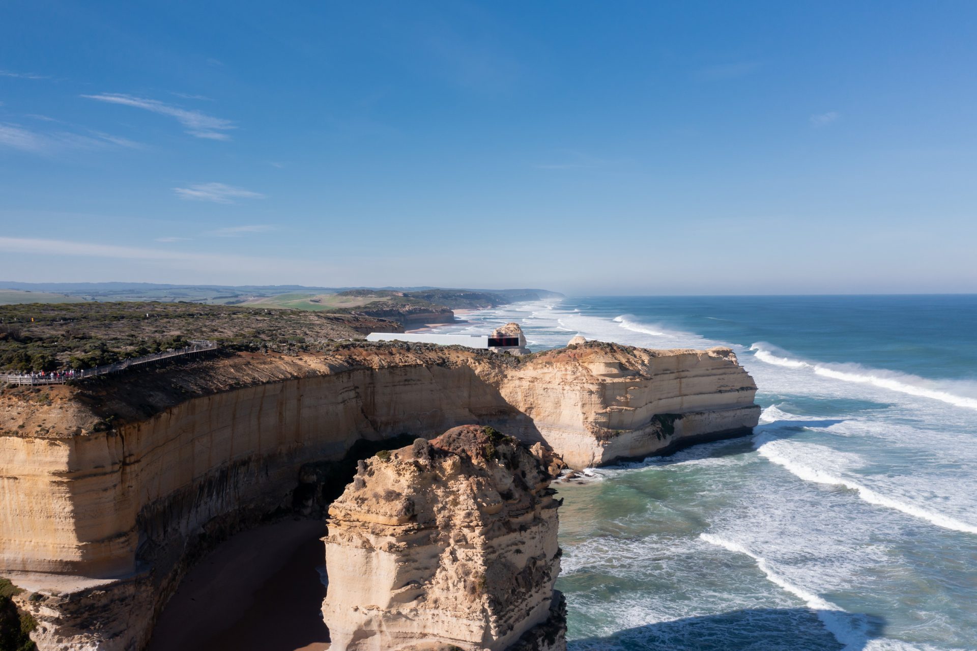 A scenic coastline view featuring rugged, towering limestone cliffs along the ocean. Waves crash against the base of the cliffs, and the coastline extends into the distance under a clear blue sky. From the Twelve Apostles lookout, greenery is visible atop the cliffs in the background.