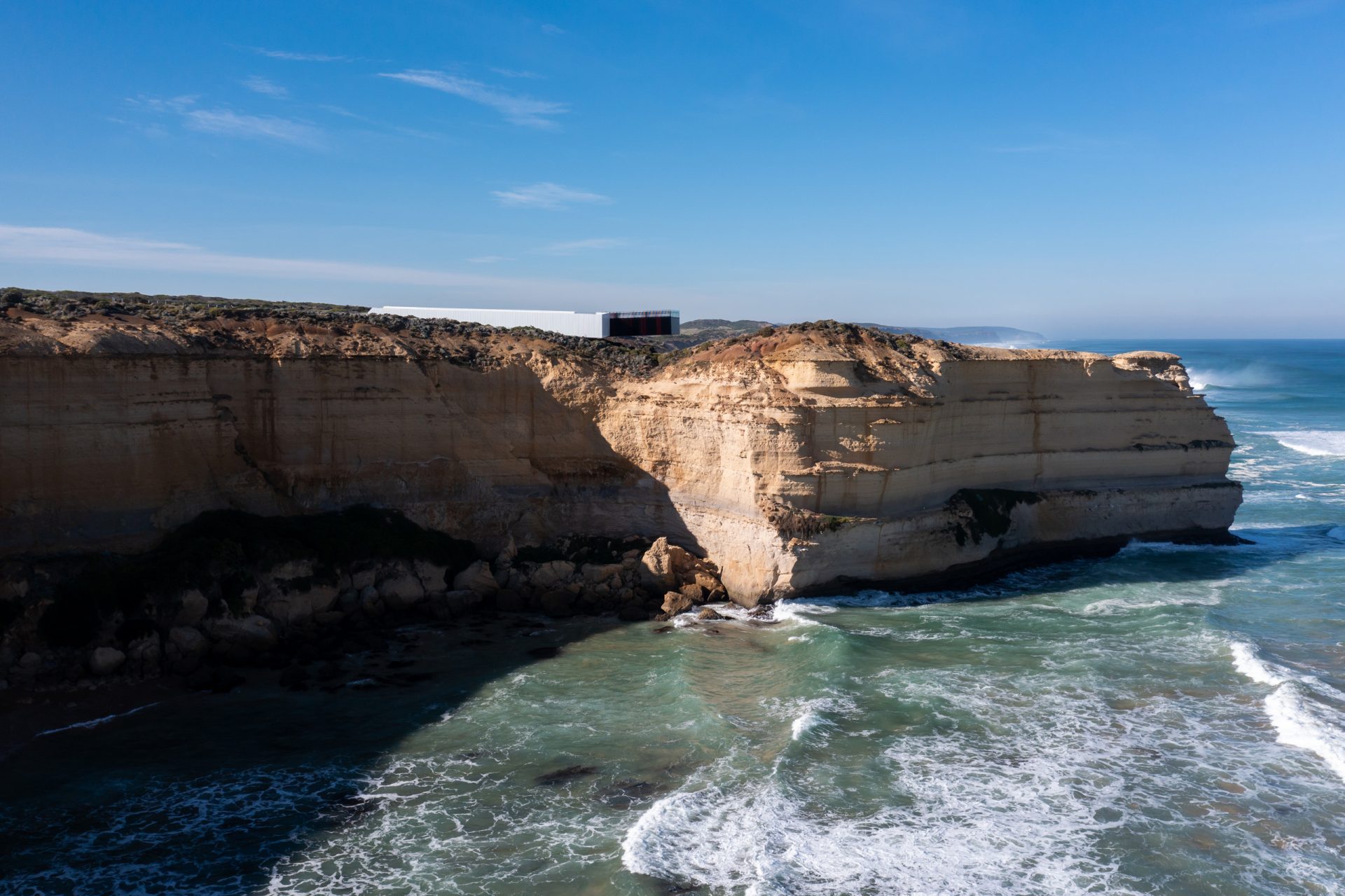 A rugged Australian coastline with golden limestone cliffs stands against an expansive blue ocean with white-capped waves. Atop the cliffs, a modern lookout with large windows overlooks the sea, set against a clear sky with scattered clouds, not far from the famed Twelve Apostles.