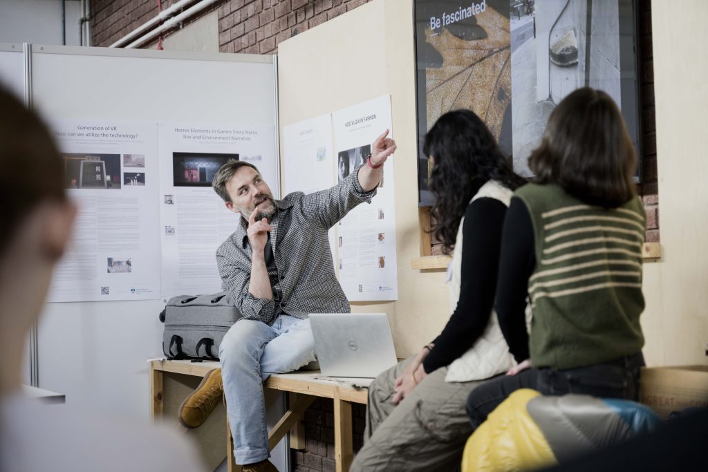 A man with a microphone points to a poster on a wall while seated on a bench, engaging with two women who are attentively listening. The setting appears to be an indoor event, perhaps organized by the National Saturday Club, with informational posters about inspiring future landscape architects displayed behind them.