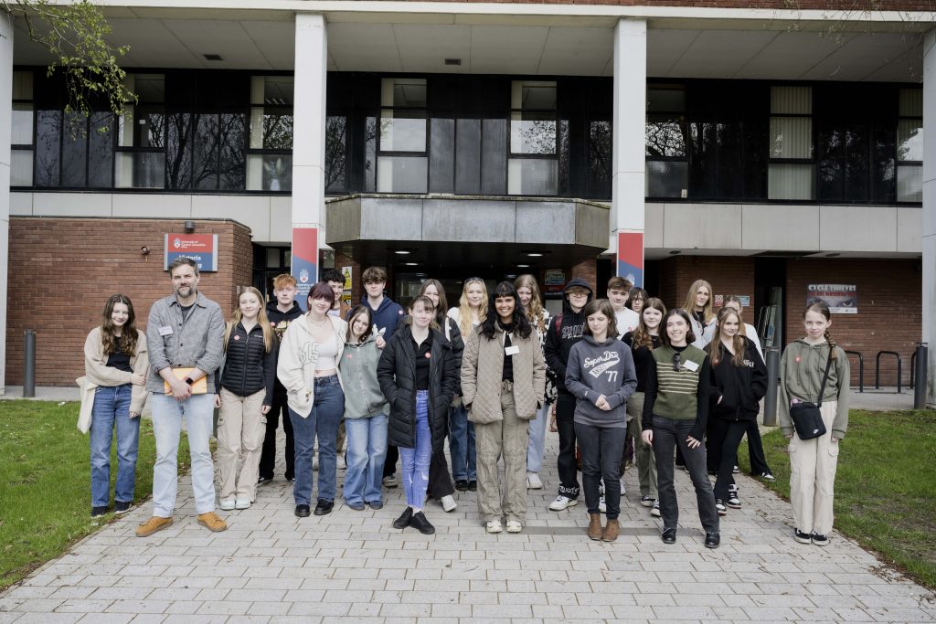 A group of people stands outside a building with a brick facade and large windows. Some are smiling, others have neutral expressions. The diverse individuals in casual attire are part of the National Saturday Club. The background features signs and noticeable greenery on the lawn.