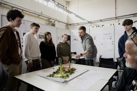 A group of young future landscape architects stands around a table with a model or project on it, while an older man from the National Saturday Club explains something and gestures towards the model. They are in a well-lit room with inspiring posters and diagrams on the walls.