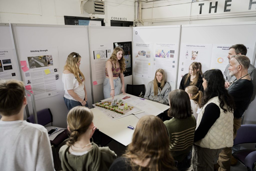 A group of people gathers around a table displaying a detailed model and some papers. The event, organized by the National Saturday Club, appears educational with posters and informational boards on the walls. Some individuals are standing while others are seated, engaged in inspiring discussions about becoming future landscape architects.