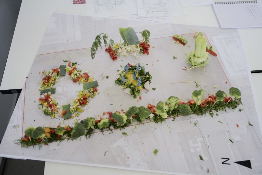 An architectural model made from vegetables and herbs is set on a white table covered with a blueprint. Created by future landscape architects, various vegetables, including broccoli, lettuce, celery, and tomatoes, are used to craft inspiring miniature representations of buildings and green spaces.