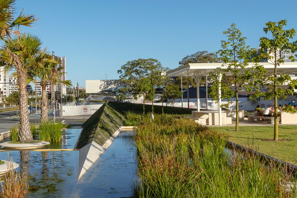 An urban park with a reflective water feature, surrounded by lush greenery and palm trees, embodies sustainability. To the right, people sit under a modern white pavilion with a partially shaded lawn area. Buildings and a clear blue sky are visible in the background.