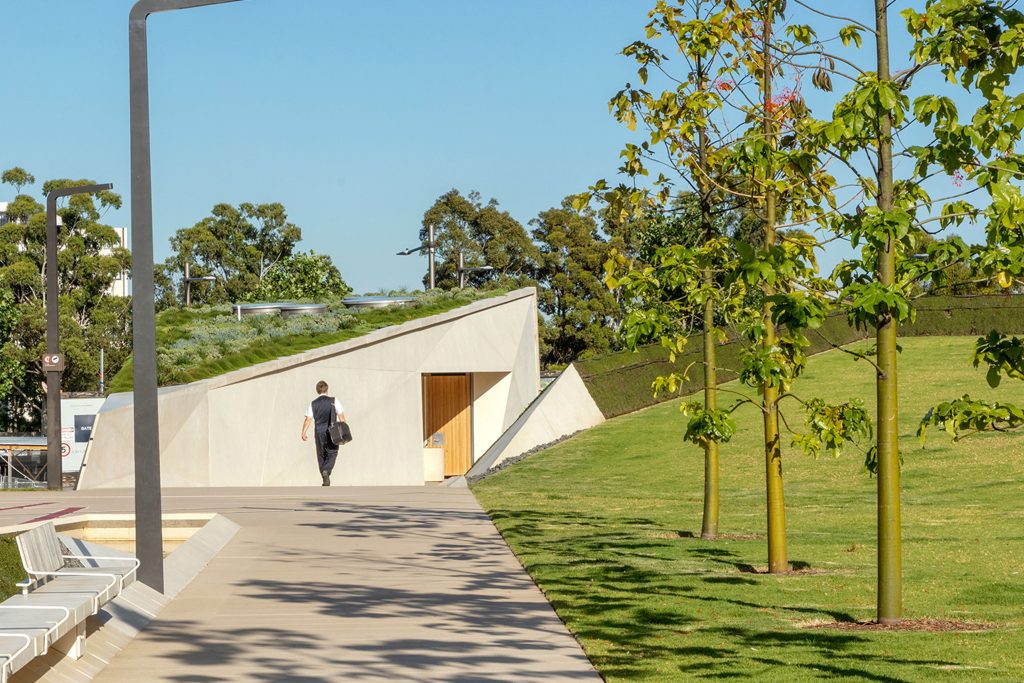 A person walks along a paved path next to a landscaped building with a green roof and recessed entrance. The modern structure, which won the Green Good Design Award for its sustainability, is set against a backdrop of trees and a clear blue sky. Young trees line the grassy area adjacent to the path.