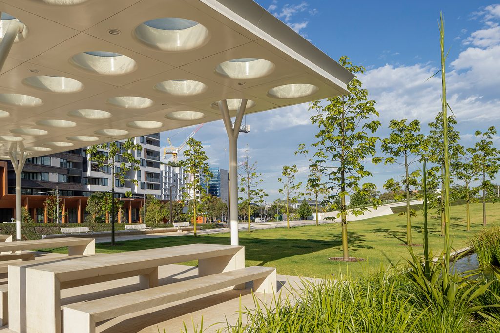 A modern park, known as Drying Green, features a covered concrete picnic area with circular gaps in the roof providing natural light. Surrounded by green lawns, young trees, and contemporary apartment buildings under a bright blue sky, this park epitomizes sustainability and has been honored with the 2024 Green Good Design Award.