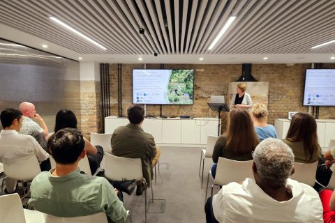 A speaker stands at a podium presenting to an audience in a modern conference room in London. The audience is seated and facing two large screens displaying slides on water quality. The room has a brick wall and a wooden slat ceiling with recessed lighting, adding to the professional atmosphere of the CIWEM event.