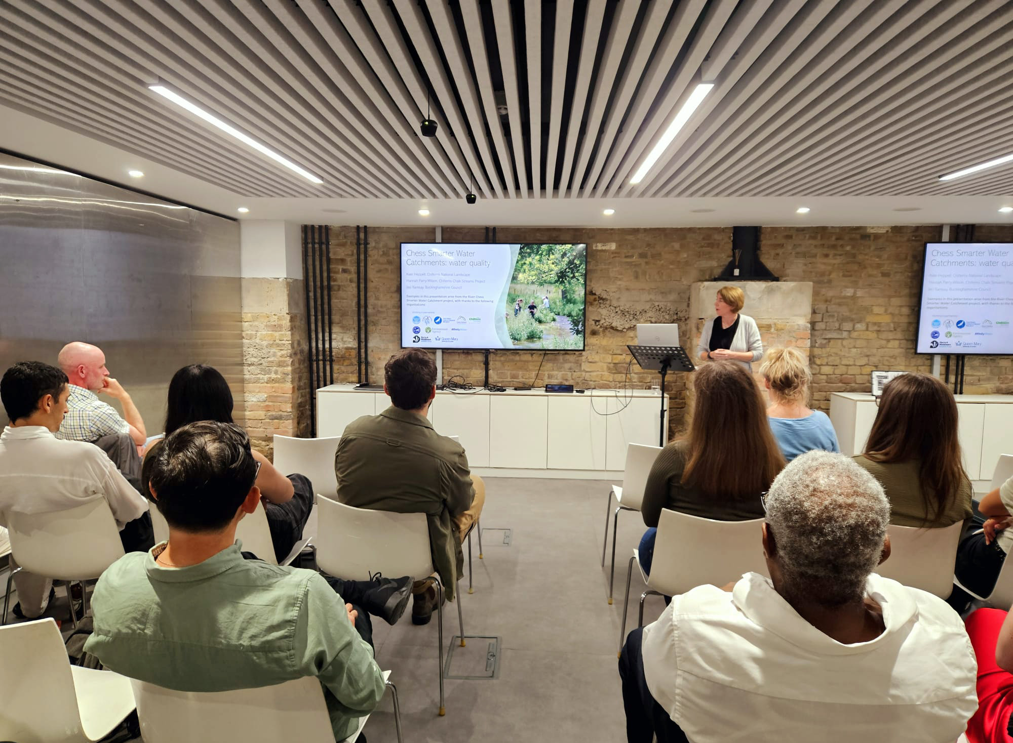 A speaker stands at a podium presenting to an audience in a modern conference room in London. The audience is seated and facing two large screens displaying slides on water quality. The room has a brick wall and a wooden slat ceiling with recessed lighting, adding to the professional atmosphere of the CIWEM event.