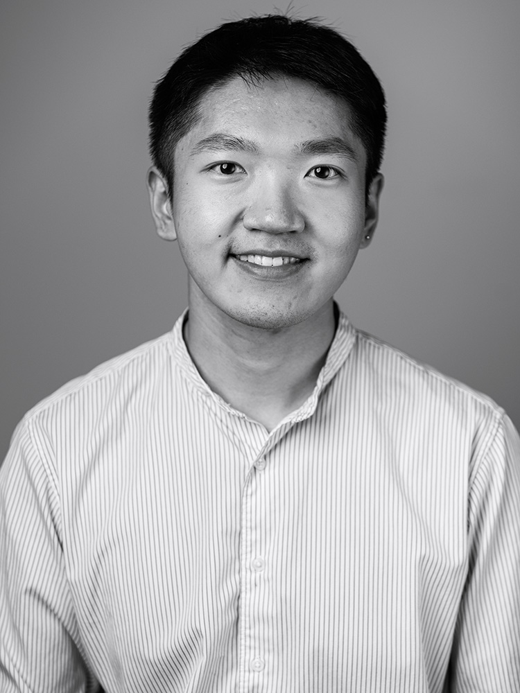 Black and white portrait of a young man with short hair, smiling at the camera. He is wearing a light-colored, vertically striped, button-down shirt with a stand-up collar designed by Yaoyao Zhang. The background is plain and neutral.