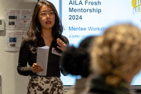 A woman is standing and speaking while holding a sheet of paper in front of a screen displaying the text "AILA Fresh Victoria 2024 Mentee Workshop." She has long hair and is wearing glasses and a black top with white patterned pants. A blurred audience is in the foreground.