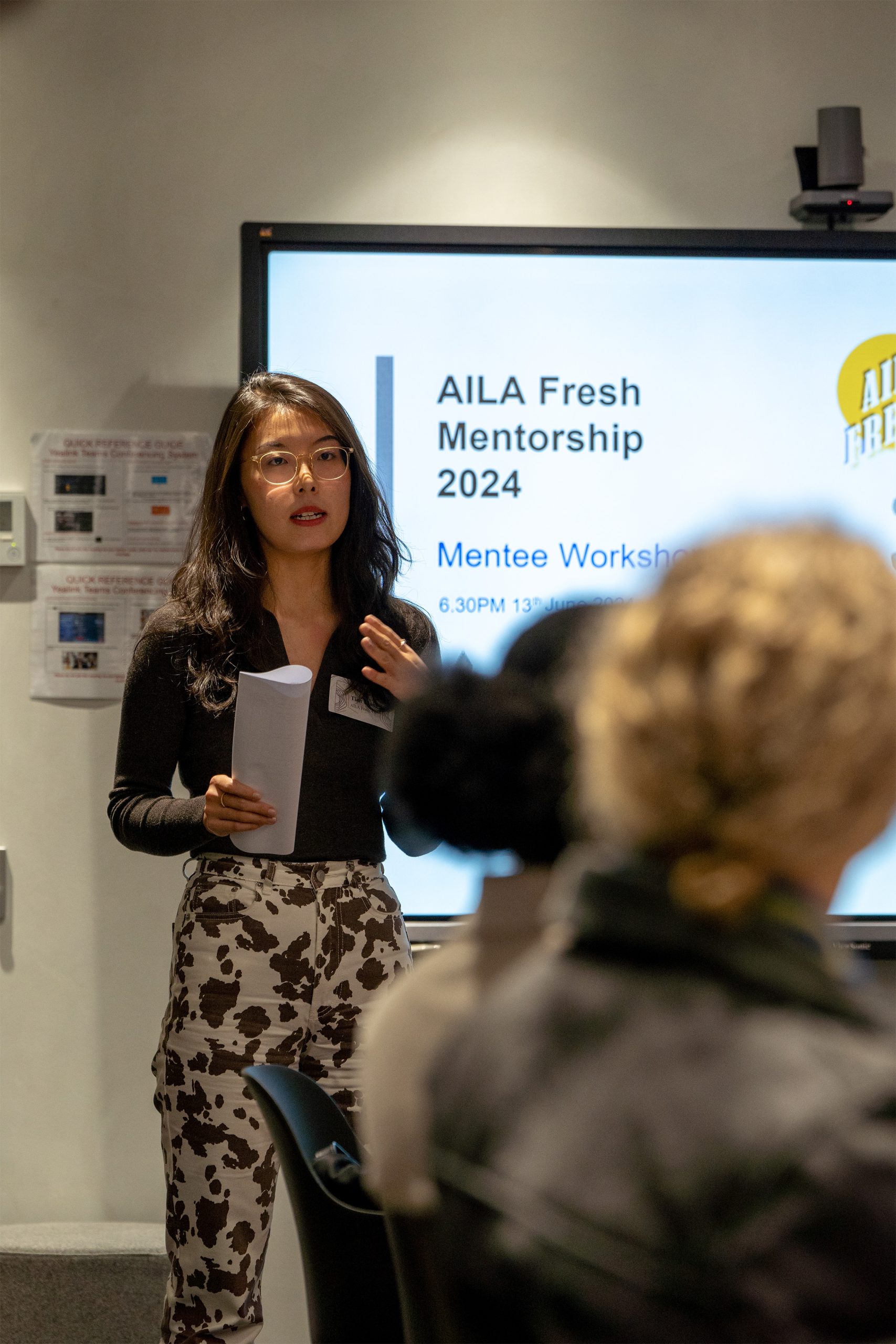 A woman is standing and speaking while holding a sheet of paper in front of a screen displaying the text "AILA Fresh Victoria 2024 Mentee Workshop." She has long hair and is wearing glasses and a black top with white patterned pants. A blurred audience is in the foreground.