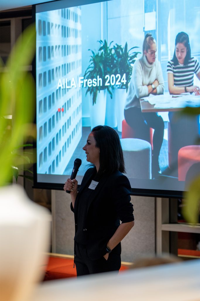 A person stands and speaks into a microphone in front of a large screen displaying "AILA Fresh Victoria." In the background, the screen shows an image of two individuals sitting and working together at a table with plants visible in the foreground, marking the vibrant launch of AILA Fresh 2024.