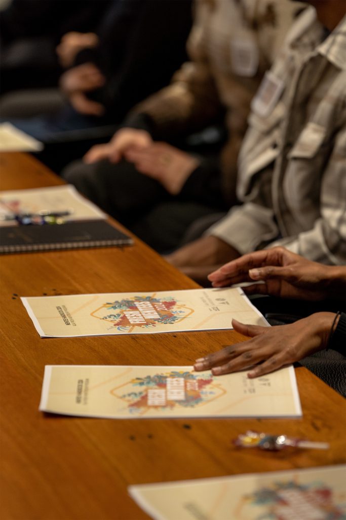 A close-up of people seated at a wooden table with printed materials in front of them. The focus is on hands resting on colorful, illustrated documents labeled "AILA 2024." The scene suggests a meeting or workshop setting. Other items, such as Fresh Victoria notebooks and small candies, are visible on the table.