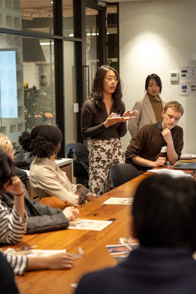 A woman stands and speaks passionately at a conference table during a meeting. She gestures with her hands as several people seated around the table listen attentively. The room, featuring a modern design with glass walls and framed articles, buzzes with excitement as AILA Fresh Victoria launches 2024.