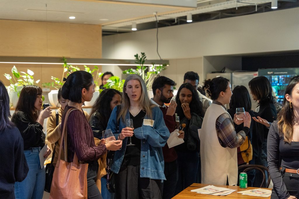 A diverse group of people gather in a modern, well-lit indoor space. Some are engaged in conversation while holding drinks, while others are standing and listening. Fresh Victoria plants decorate the background and 2024 event papers on a table in the foreground add to the ambiance.