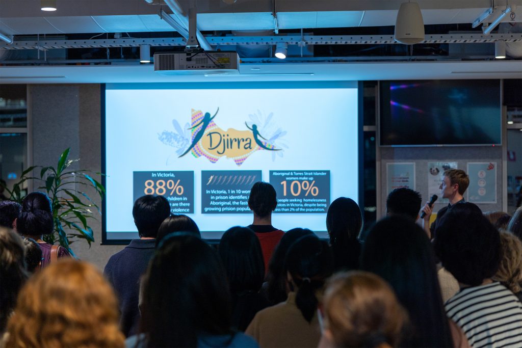 A crowd of people watches a presentation in a dimly lit room. The slide on the screen displays the text "Djirra" and statistics about imprisonment rates, starting with "88% in Victoria" and "10% in Aboriginal & Torres Strait Islander". AILA's 2024 speaker stands to the right, discussing Fresh Victoria initiatives.