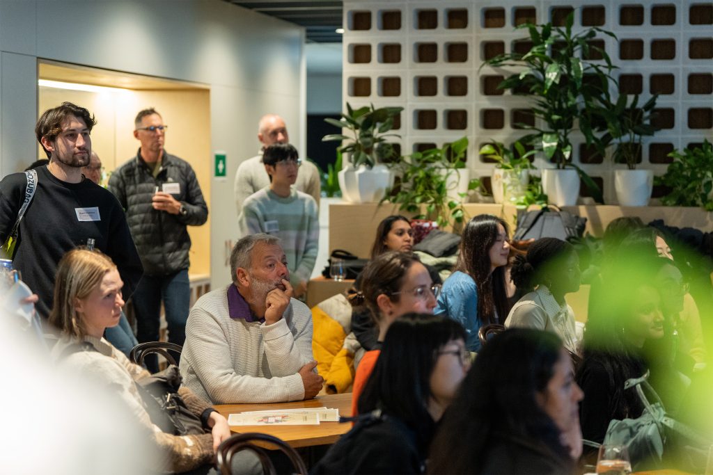 A diverse group of people, including Victoria, seated and standing in a modern, plant-filled room attentively watching a presentation in 2024. Some are taking notes or sipping drinks, and the atmosphere appears engaged and focused as they discuss AILA's innovations.