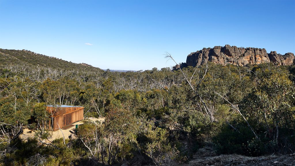 A modern cabin with a large window is situated amidst dense greenery in a rugged, rocky landscape. The rocky escarpment rises prominently in the background under a clear blue sky. This exemplary piece of architecture has earned accolades and features in the International Architecture Awards, blending harmoniously with its natural surroundings.