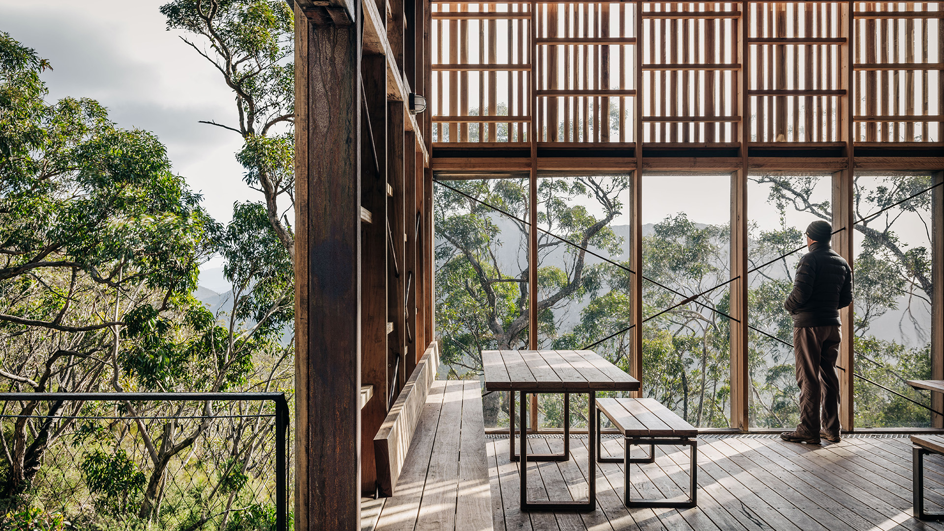 A person stands inside a wooden structure with large windows, looking out at a lush, green forest. The space, honored with dual accolades in this year's International Architecture Awards, features wooden tables and benches. Sunlight filters through the trees, creating a serene, natural atmosphere.