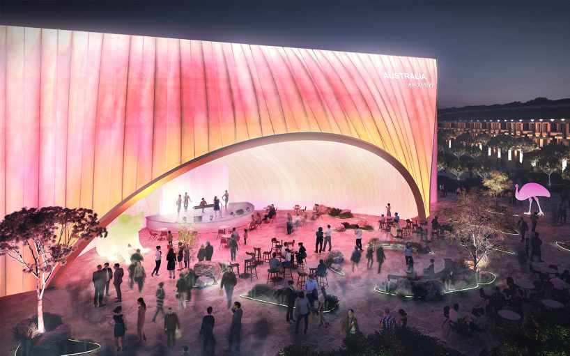 Nighttime scene of the Australian Pavilion at Osaka Expo 2025, featuring a large, illuminated wave-like structure with "Australia" on it. People are gathered around chatting and dining, with mist around their feet and a glowing pink flamingo light in the distance.