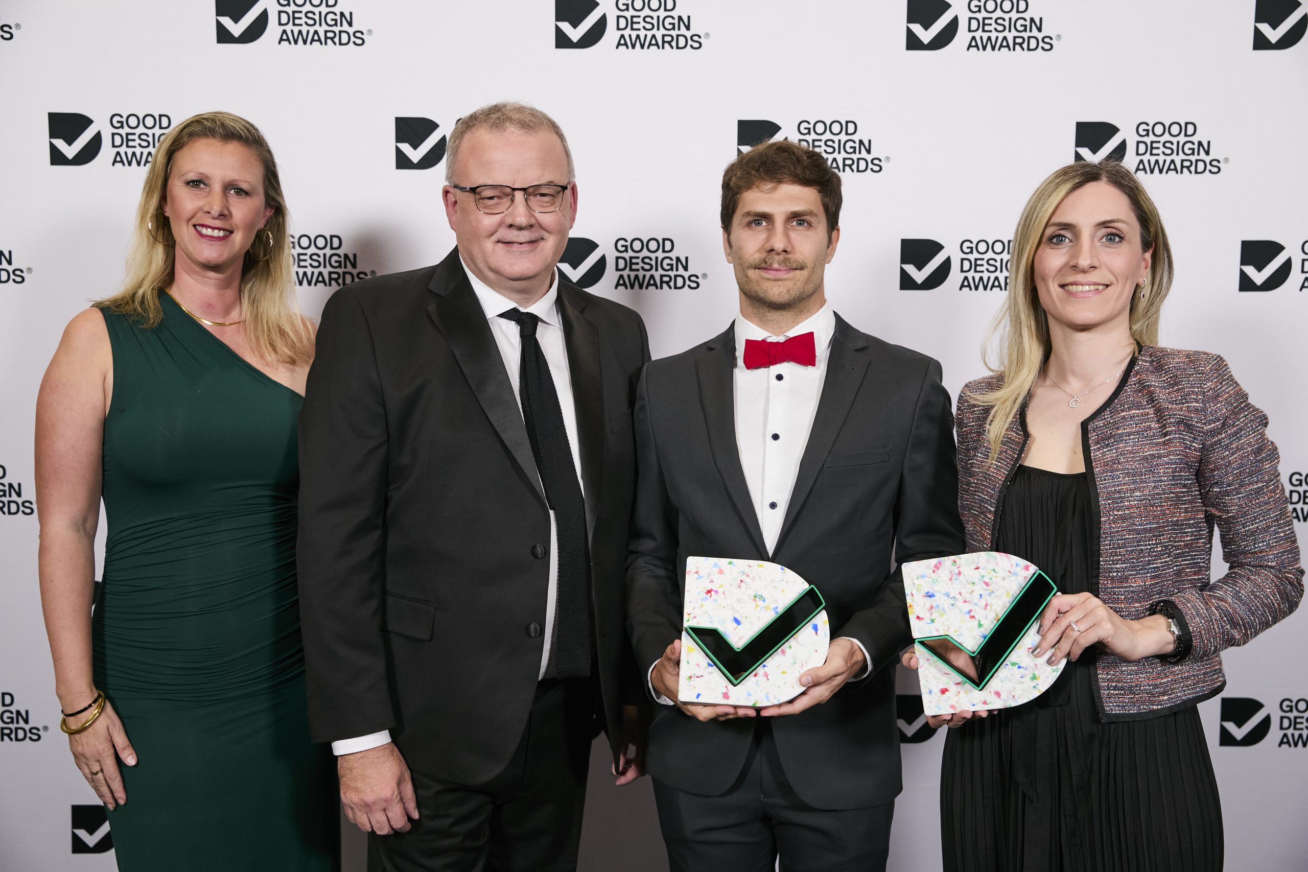 Four people are posing for a photo at the Good Design Awards. Two individuals in the center are proudly holding dual accolades shaped like triangles with a green checkmark design. The backdrop shows the Good Design Awards logo repeatedly. Formal attire is worn by all.