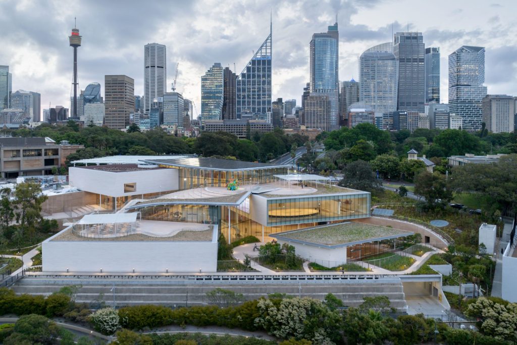 Aerial view of an art museum with modern architecture, characterized by sleek lines and large glass windows, surrounded by greenery. In the background, a cityscape with numerous skyscrapers and a prominent communications tower stands against a cloudy sky, showcasing adaptive urban design.