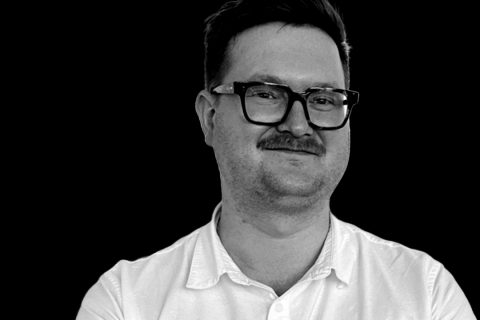 A person with short hair, glasses, and a mustache is wearing a white, collared shirt and smiling slightly. The backdrop is black, creating a high contrast with their light-colored shirt—a photo taken during the 2024 Innovation Symposium featuring Thomas Chapman.