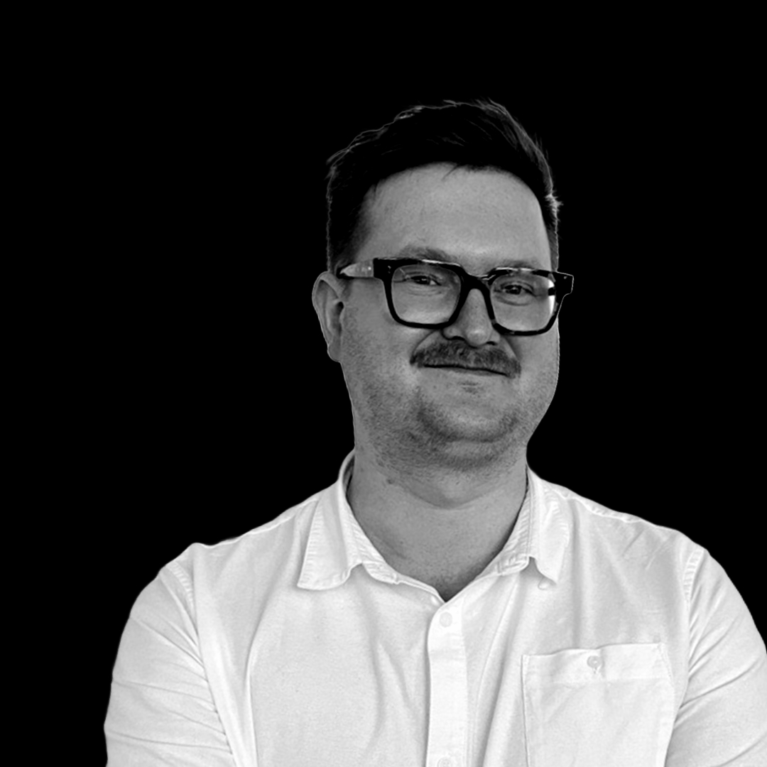 A person with short hair, glasses, and a mustache is wearing a white, collared shirt and smiling slightly. The backdrop is black, creating a high contrast with their light-colored shirt—a photo taken during the 2024 Innovation Symposium featuring Thomas Chapman.