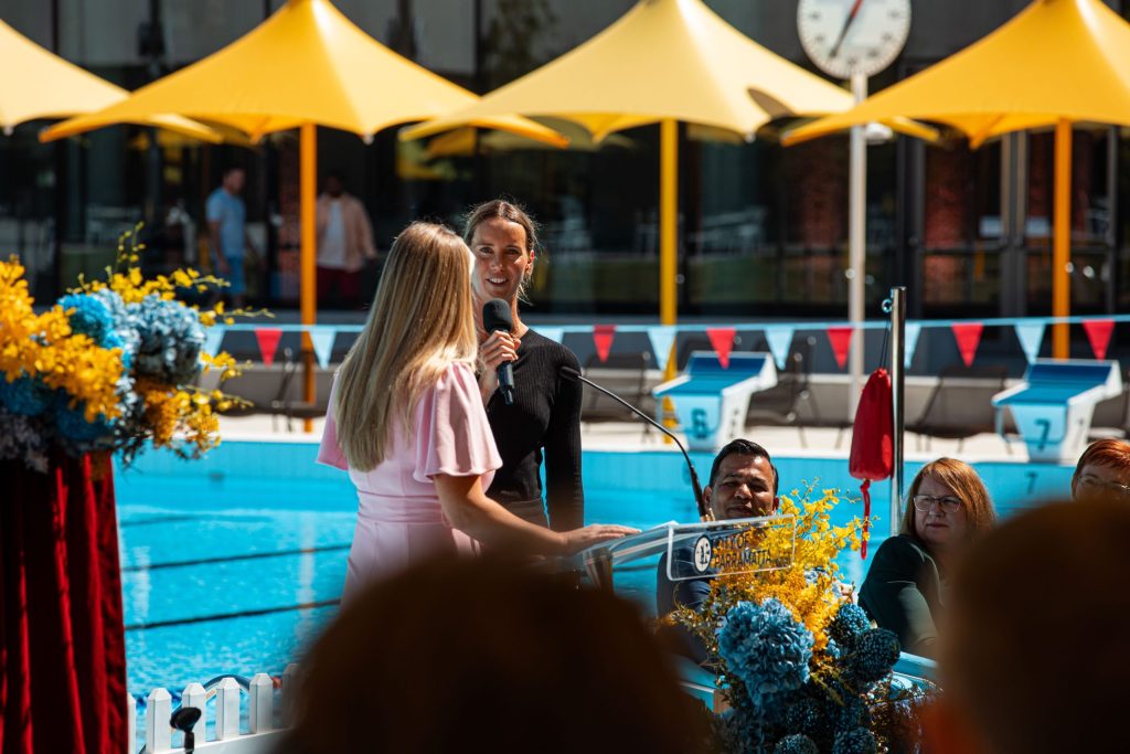 At the Parramatta Aquatic Centre, two women stand by a podium at a swimming pool event, with yellow umbrellas adding a splash of color. One wears a pink dress and the other, in black, holds a microphone. A decorative floral arrangement adorns the left as attendees enjoy this moment on the world stage.