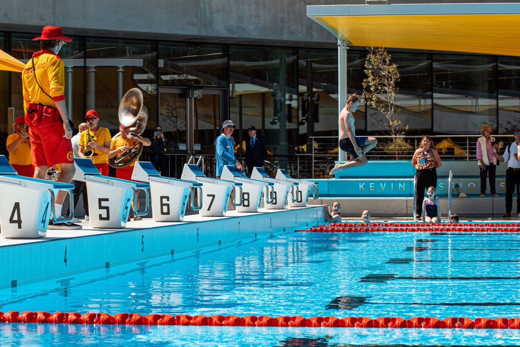 A brass band performs beside an outdoor swimming pool at Parramatta Aquatic Centre. A boy makes a big splash as he jumps into the clear blue water, delighting spectators. The brightly lit pool area is lively, resembling a world stage and creating a festive atmosphere.